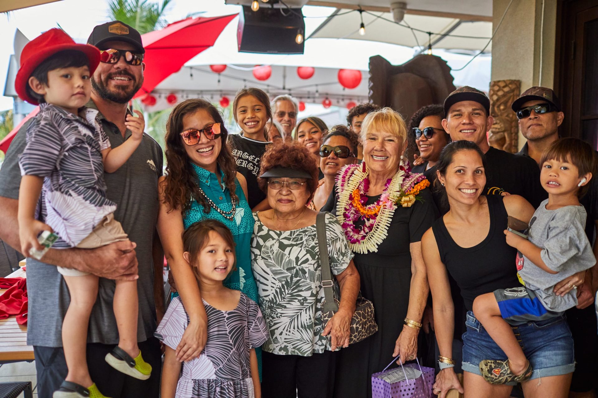 A large, multi-generational local family arriving at Tiki's Grill & Bar to celebrate an occasion, some members wearing lei, showcasing the restaurant's ability to host large groups.
